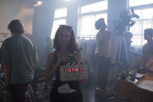 Maura on set of her film Disoriented, holding a clapper while the crew sets up behind her on set.